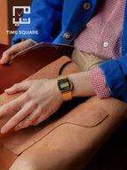 A person wearing a gold-tone digital watch with a square face and gold link bracelet, resting on a tan leather surface. The person is dressed in a blue blazer over a red-and-white gingham shirt, with a visible TIME SQUARE logo in the upper left corner.