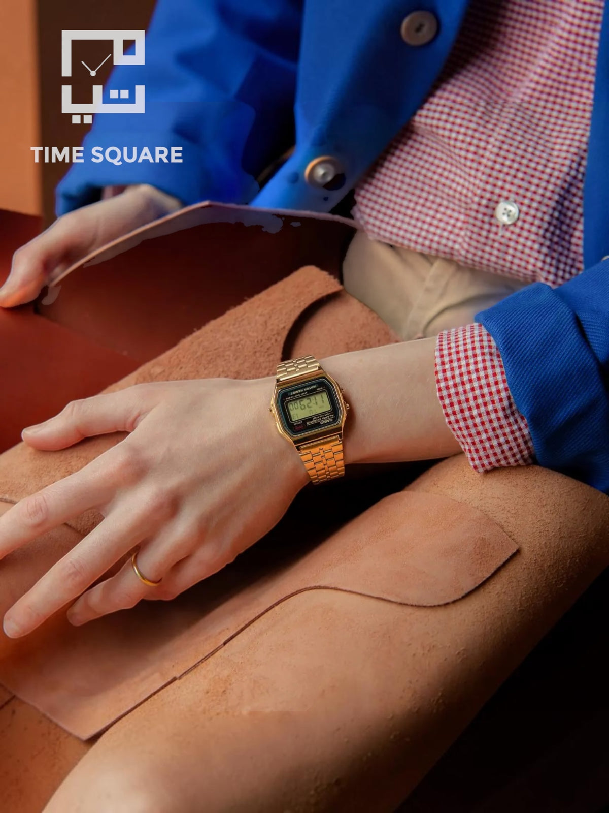 A person wearing a gold-tone digital watch with a square face and gold link bracelet, resting on a tan leather surface. The person is dressed in a blue blazer over a red-and-white gingham shirt, with a visible TIME SQUARE logo in the upper left corner.