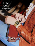 A person wearing a gold-tone digital watch with a black face and textured strap, seated in a warm-toned interior. The person is dressed in a brown leather jacket and blue jeans, holding a brown leather clutch.