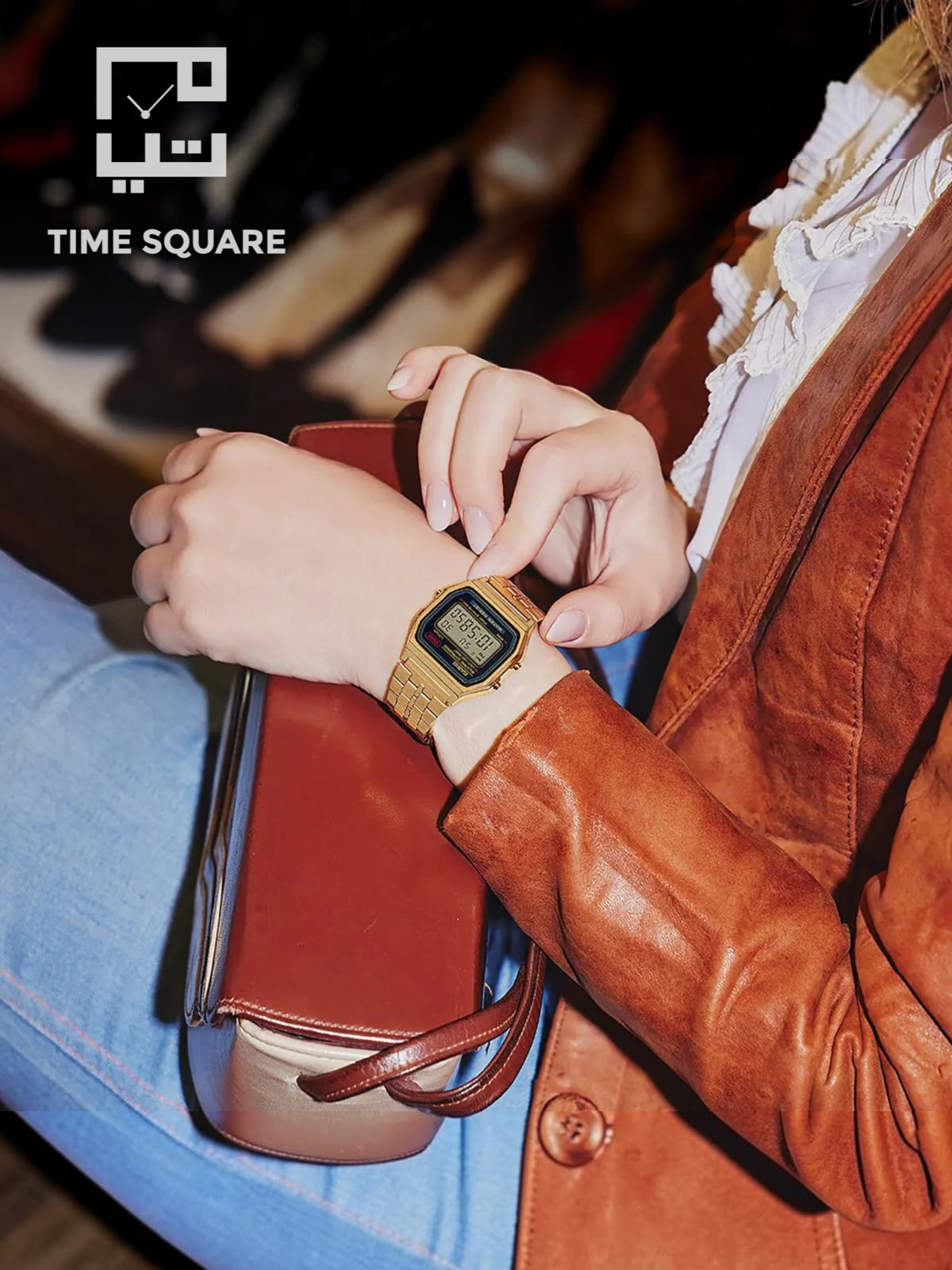 A person wearing a gold-tone digital watch with a black face and textured strap, seated in a warm-toned interior. The person is dressed in a brown leather jacket and blue jeans, holding a brown leather clutch.