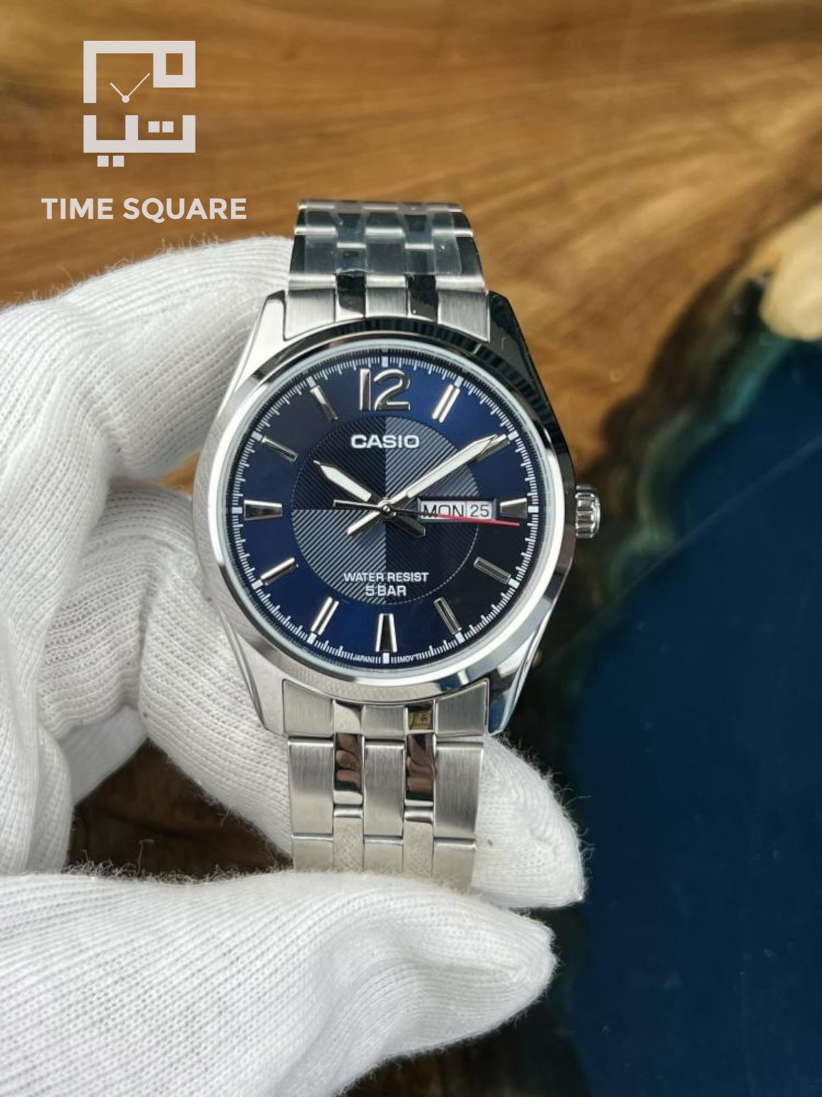 A Casio analog wristwatch with a blue dial and silver stainless steel case and link bracelet, held by a white-gloved hand against a wooden background.