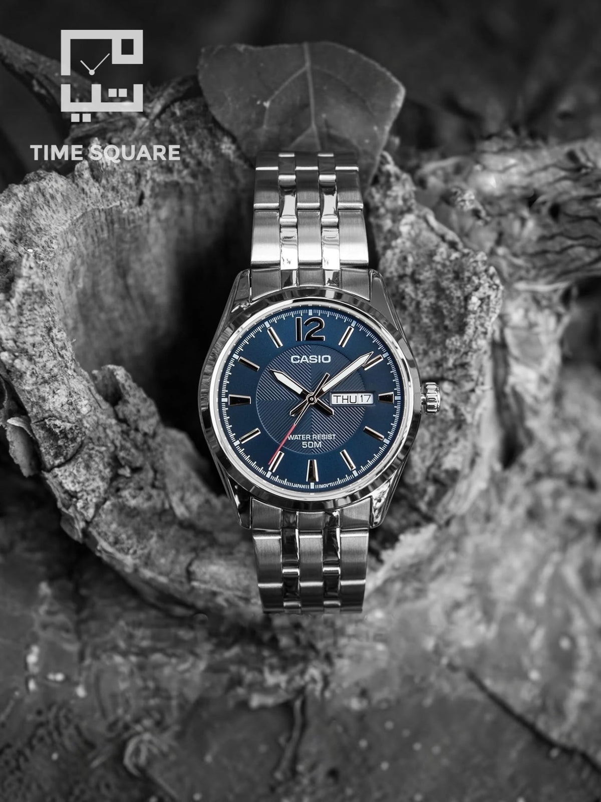 A Casio men's watch with a blue dial and silver stainless steel case and bracelet, displayed on a textured rock surface. The image is in black and white, with a 'TIME SQUARE' logo watermark in the top left corner.