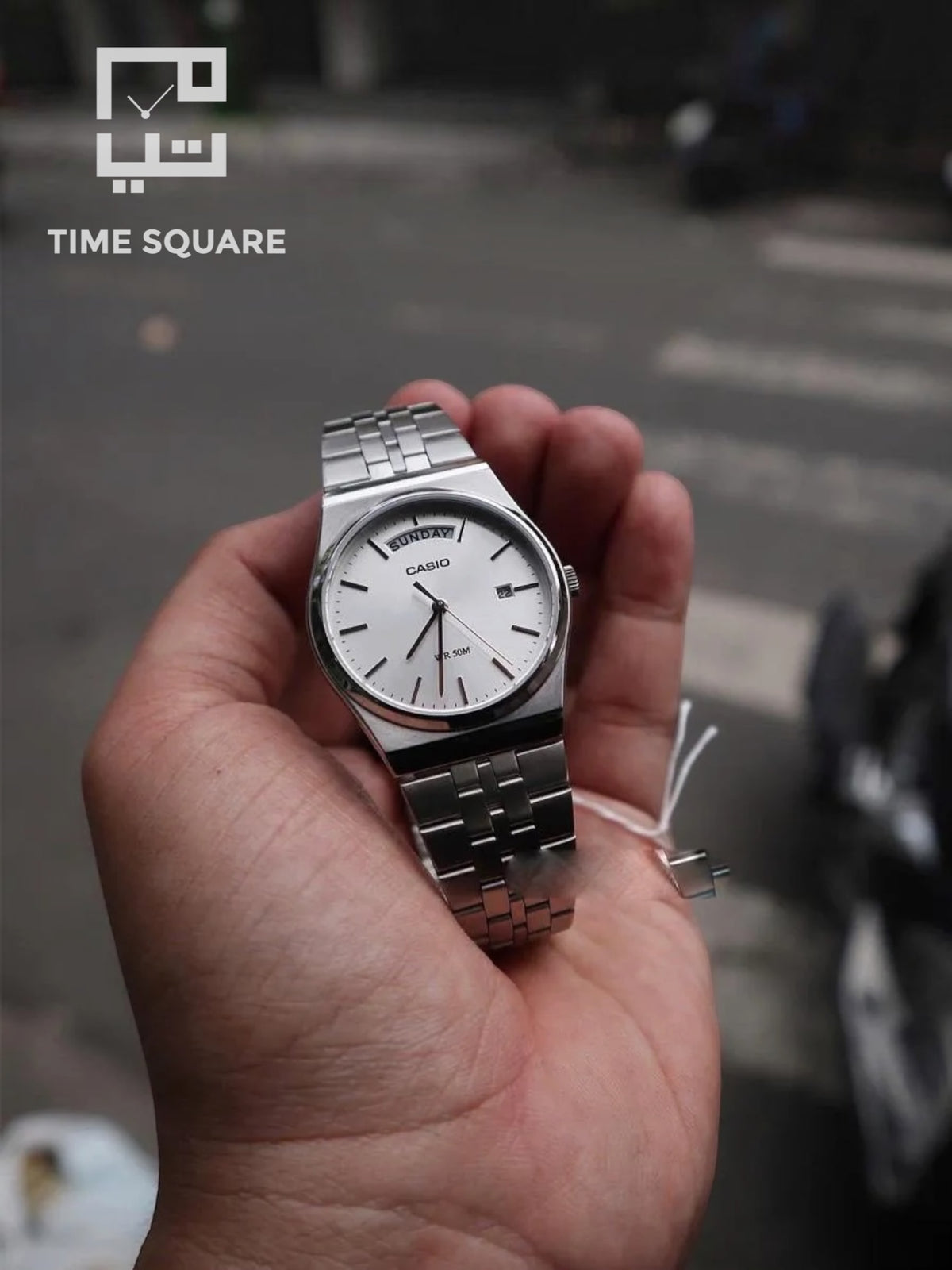 A silver Casio watch with a white dial and metal link bracelet held in a person's hand, with a blurred street background and a TIME SQUARE watermark in the top left.
