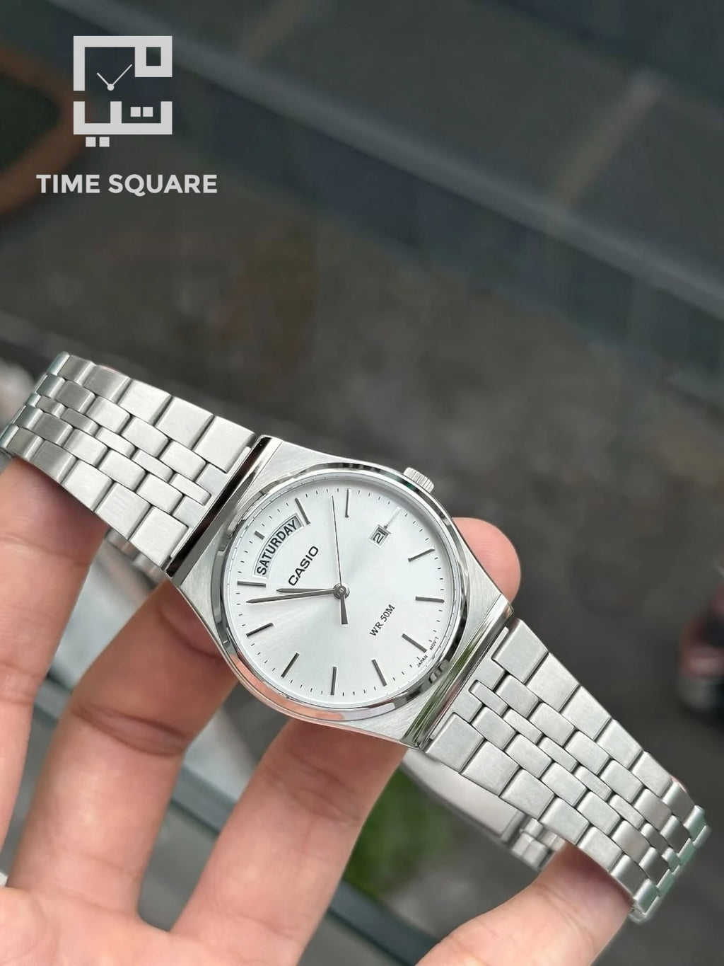 A silver Casio watch with a white dial and metal link bracelet, held in a person's hand against a blurred outdoor background. The TIME SQUARE logo is visible in the top-left corner.