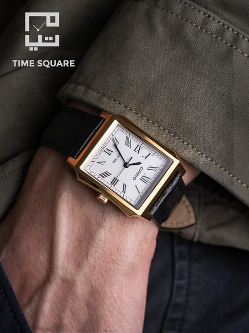 A close-up of a wrist wearing a Casio rectangular watch with a gold-tone case, white dial, and black leather strap, set against an olive green jacket and dark pants. The TIME SQUARE logo is visible in the top-left corner.