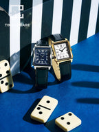 Two Cartier Tank watch models displayed on a blue surface with black and white striped background panels and cream-colored dominoes. One watch has a black dial and black strap, and the other has a white dial and gold-tone case with a black strap.