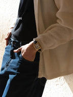 A close-up lifestyle shot of a person wearing a gold-toned rectangular watch with a dark strap, styled with a black t-shirt, dark blue jeans, and a cream-colored knit cardigan.