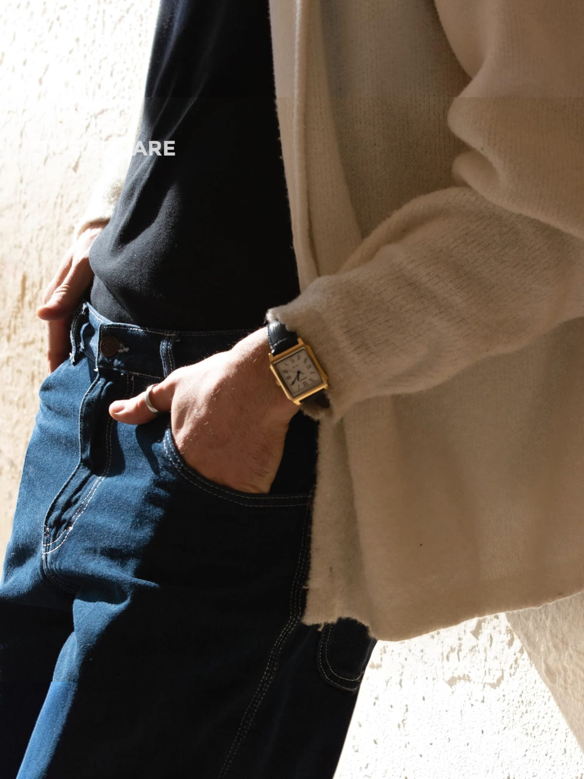 A close-up lifestyle shot of a person wearing a gold-toned rectangular watch with a dark strap, styled with a black t-shirt, dark blue jeans, and a cream-colored knit cardigan.