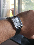 A men's rectangular analog watch with a silver-tone case, white dial, and black leather strap, shown worn on a person's wrist in a real indoor setting near a window.