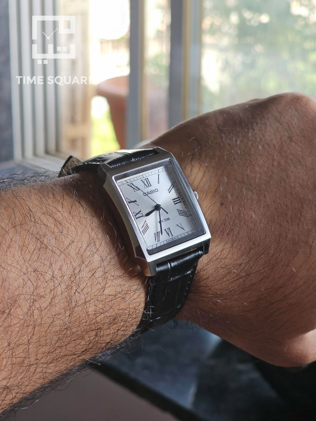 A men's rectangular analog watch with a silver-tone case, white dial, and black leather strap, shown worn on a person's wrist in a real indoor setting near a window.