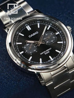 A Casio analog watch with a silver stainless steel case and bracelet, featuring a black dial with white markings and sub-dials, set against a dark blue textured background.