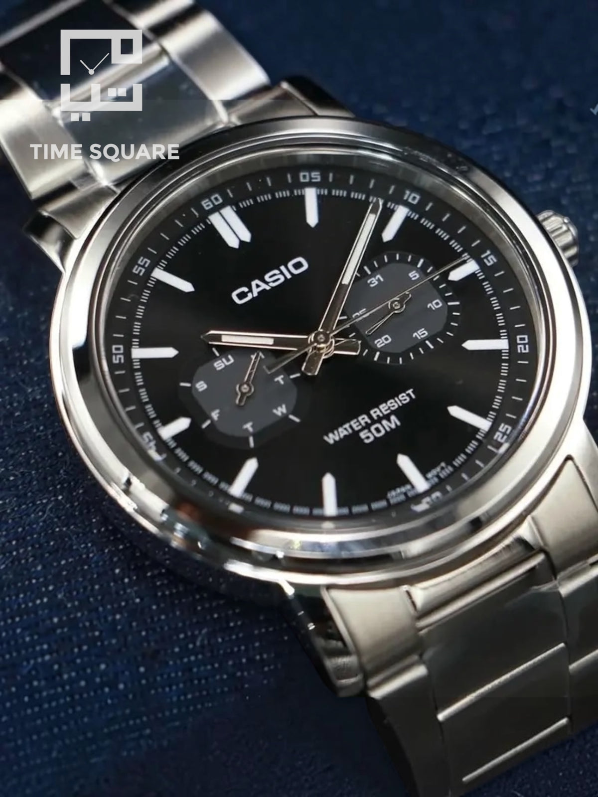 A Casio analog watch with a silver stainless steel case and bracelet, featuring a black dial with white markings and sub-dials, set against a dark blue textured background.