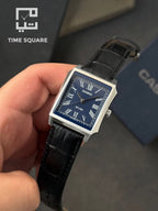 A Casio rectangular analog watch with a blue dial, silver-tone case, and black leather strap, held in a hand. The background includes a Casio box and a Time Square watermark.