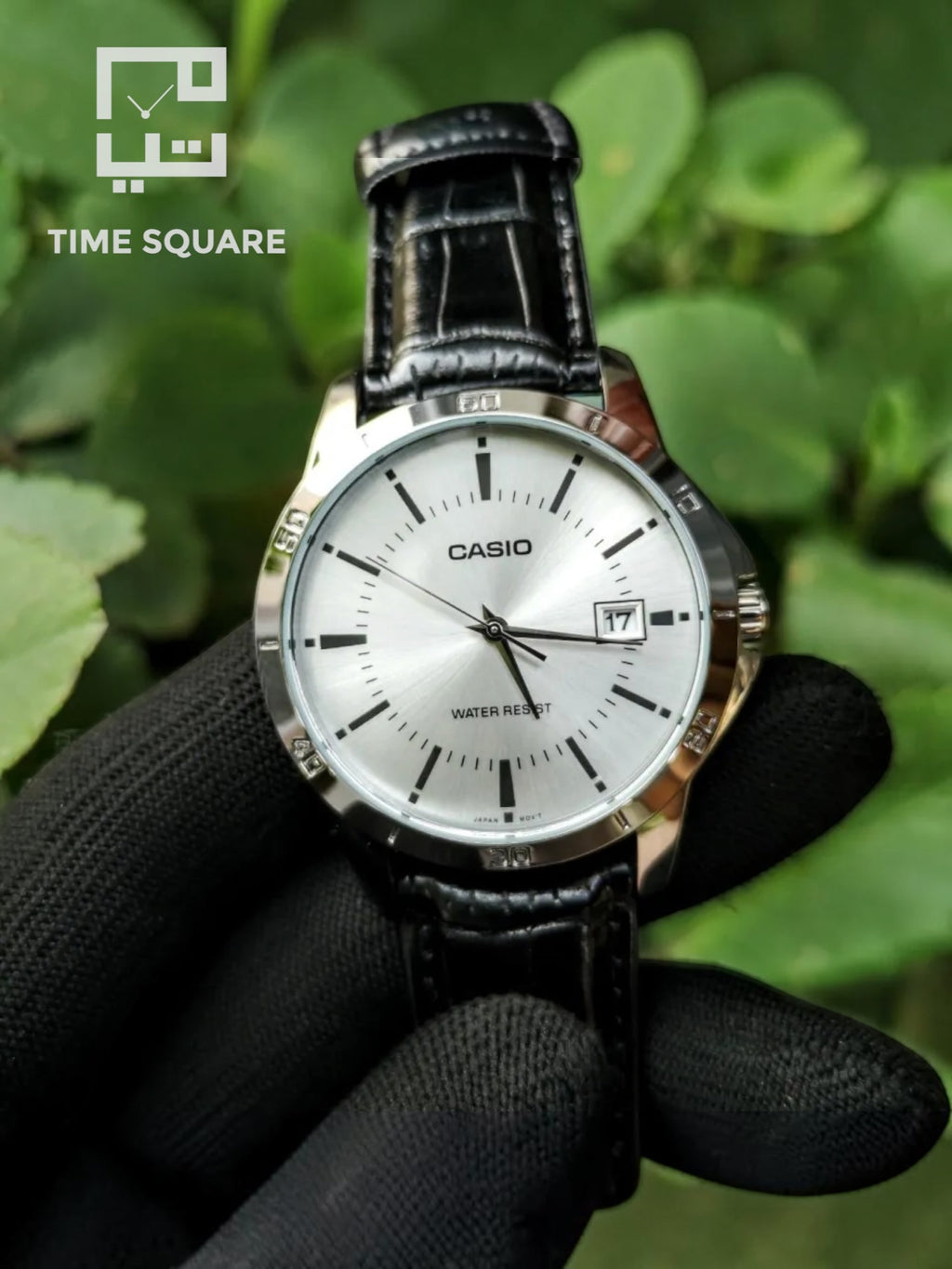 A Casio analog watch with a silver dial and black leather strap, held by a hand wearing a black textured glove against a background of green leaves. The TIME SQUARE logo is visible in the top-left corner.
