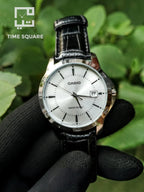 A Casio analog watch with a silver dial and black leather strap, held by a hand wearing a black textured glove against a background of green leaves. The TIME SQUARE logo is visible in the top-left corner.
